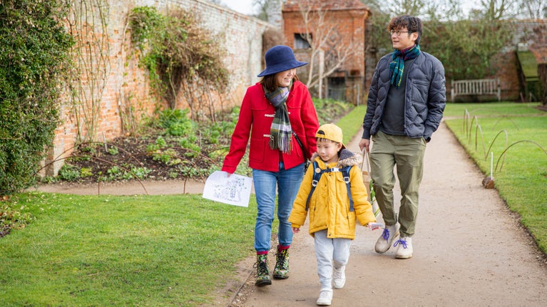 A mum is wearing a blue brimmed hat while walking down a garden path, turning and smiling to her young child - a boy wearing a bright yellow jacket, while the dad walks behind smiling and looking out across the walled garden.